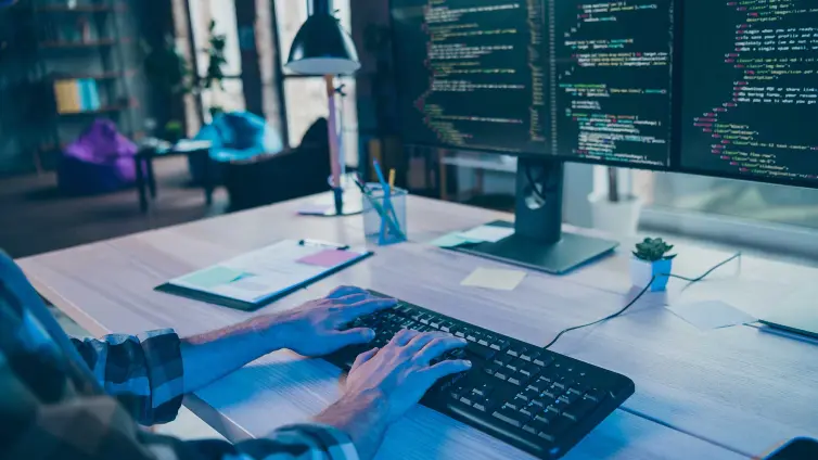 Hands typing on a keyboard in front of two monitors showing code.